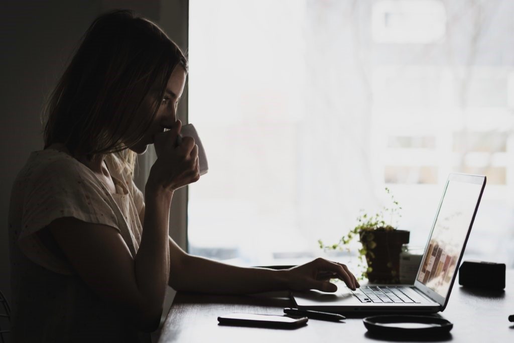 Woman drinking coffee in front of her window while looking at her computer.