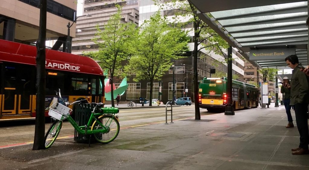 Bike share bike parked on sidewalk as buses drive by and man checks his cell phone.